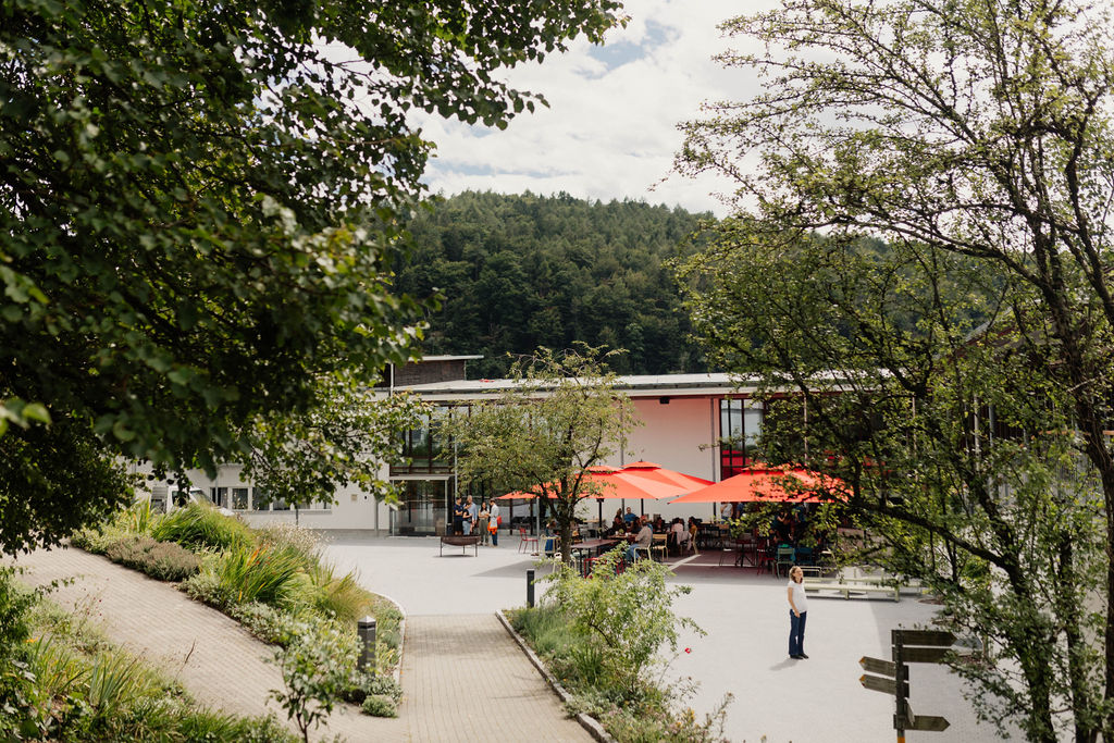 Blick auf den Hof in Arbogast, mit roten Sonnenschirmen, umrundet von Bäumen.